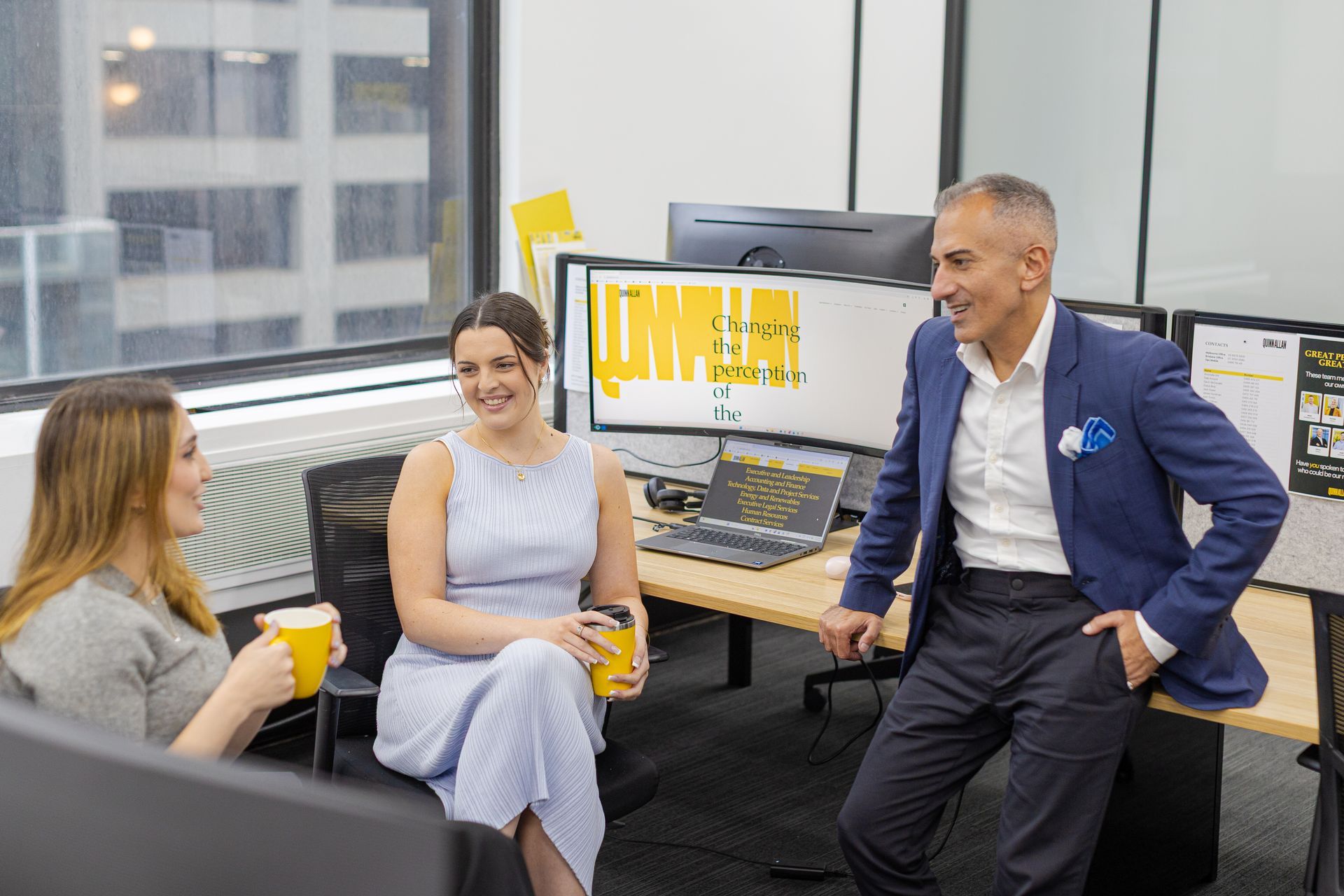 Three colleagues talking in a bright office beside a computer monitor