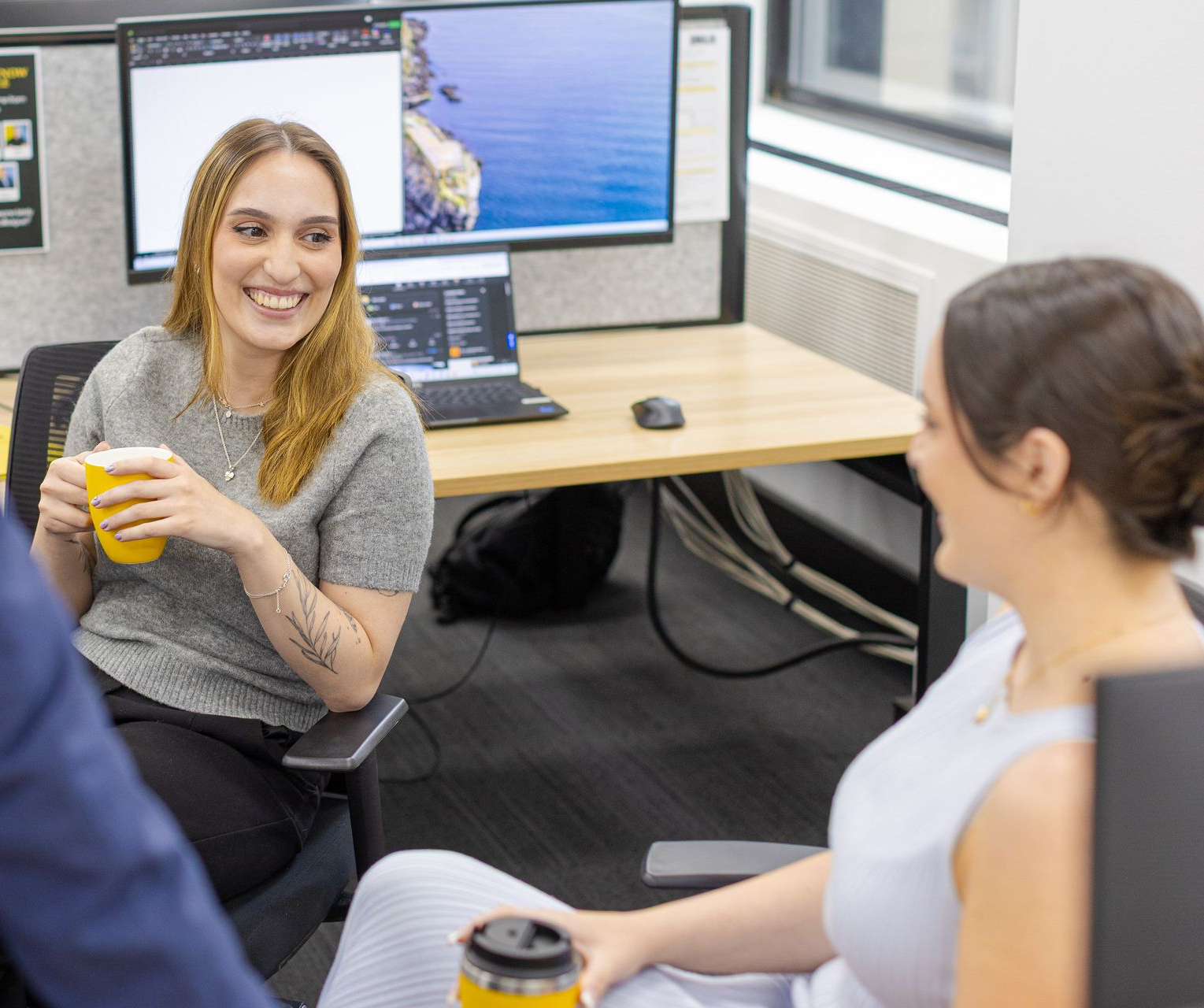 Two coworkers talking in an office, one smiling with a drink at a desk with dual monitors.