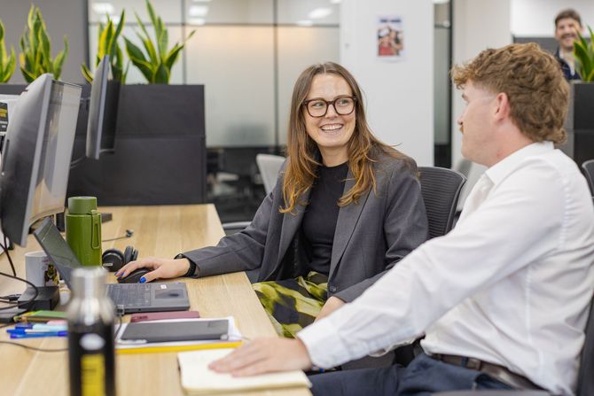 Two coworkers talking at desks in a modern office, one smiling at a computer monitor