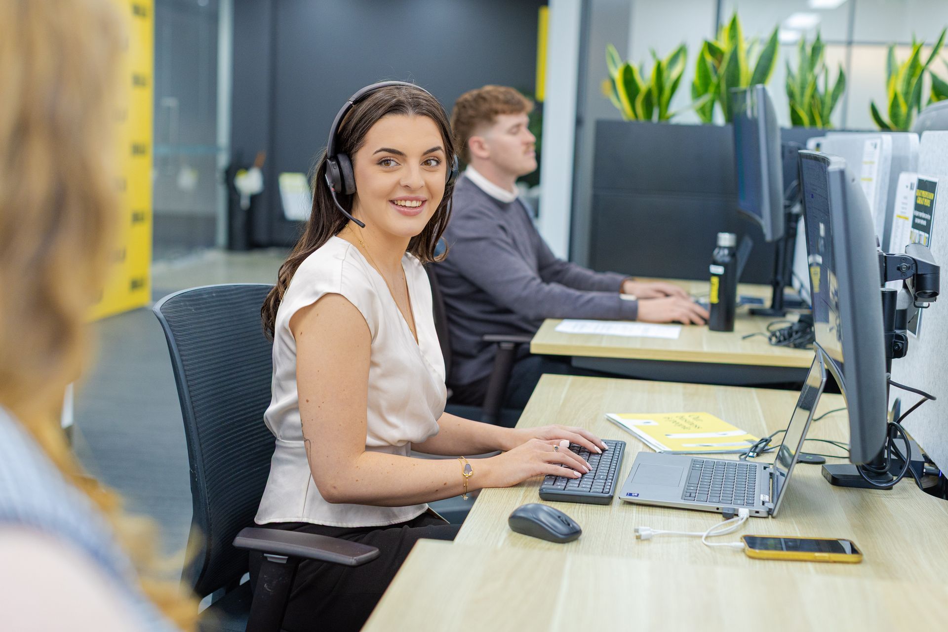 Woman working at a desk in an office, typing at a computer and smiling at the camera.