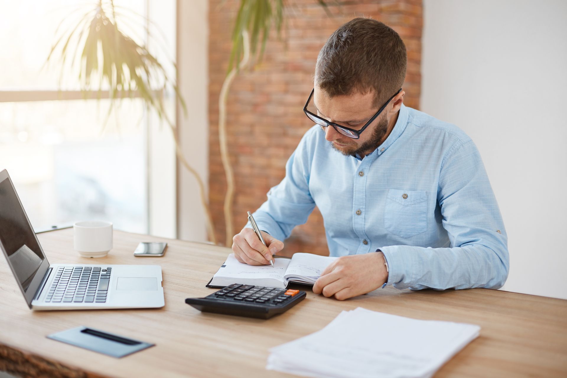 Man in blue shirt calculating with a calculator and writing on paper at a desk with a laptop