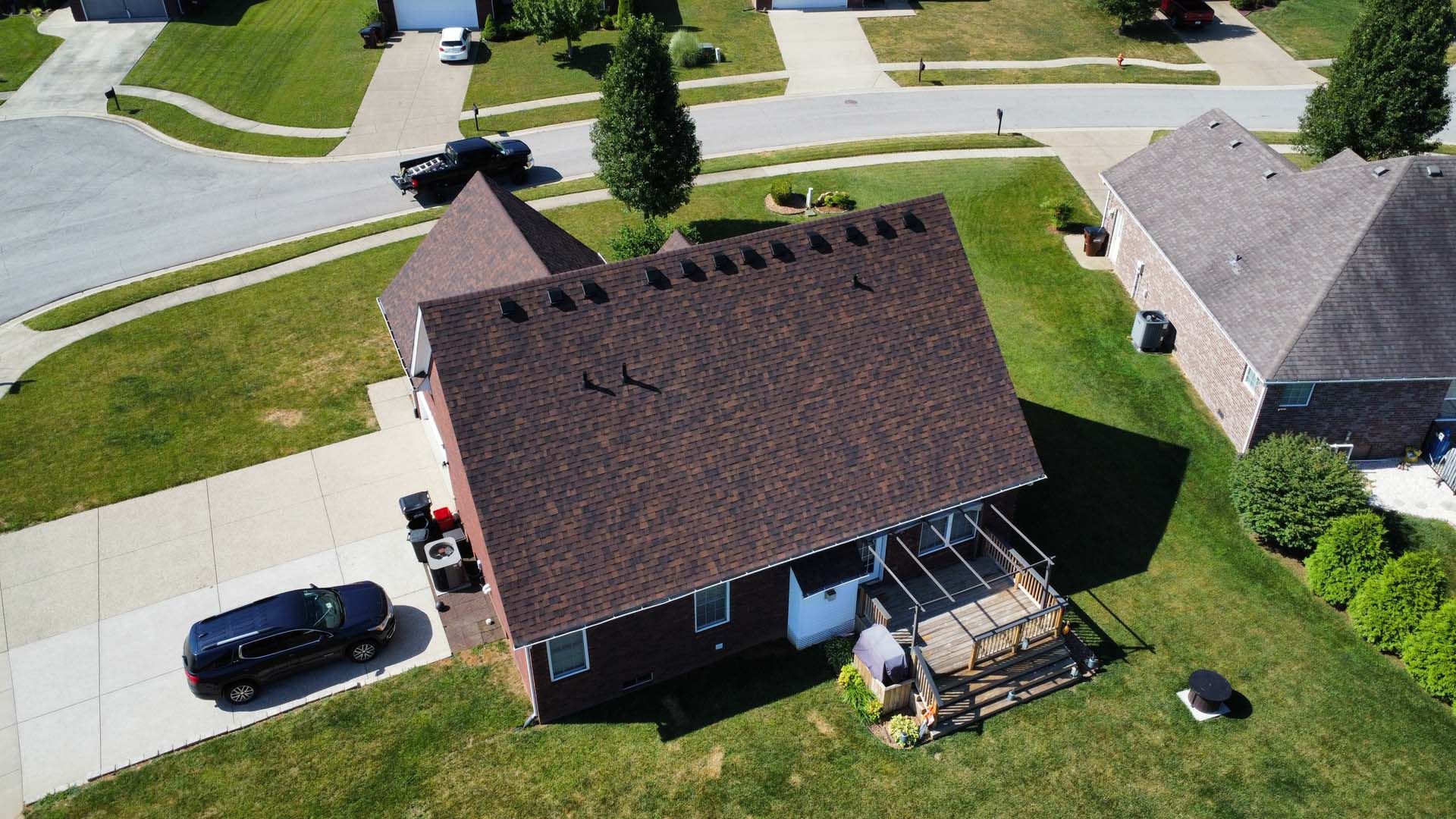 An aerial view of a single-story brick house with a brown shingled roof, a driveway, a deck, and a neighboring home.