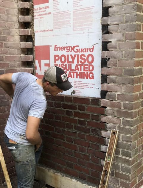 A worker in a baseball cap inspects a partially bricked-up wall opening filled with insulating board.