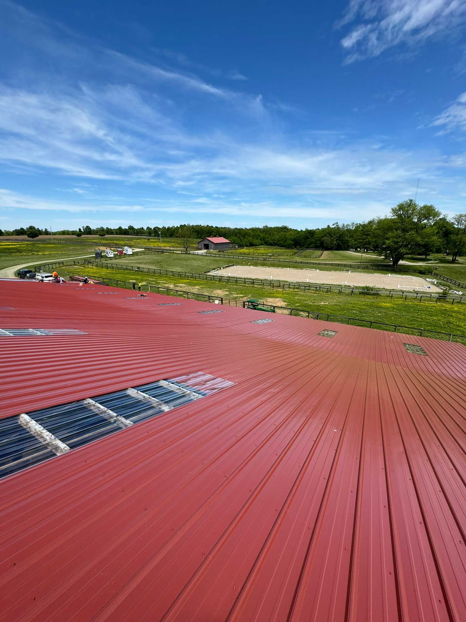 A high-angle view of a large, red metal roof with solar panels, overlooking a sunny, grassy rural landscape.