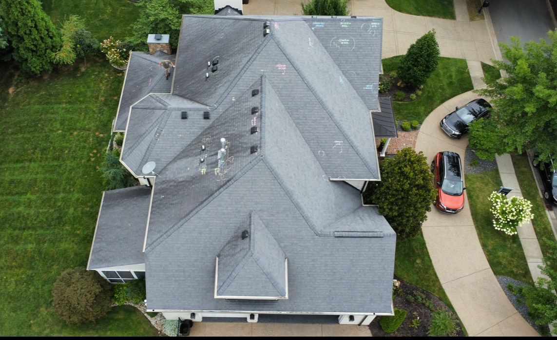 An aerial view of a gray shingled residential roof with multiple peaks, surrounded by green lawns and a paved driveway.
