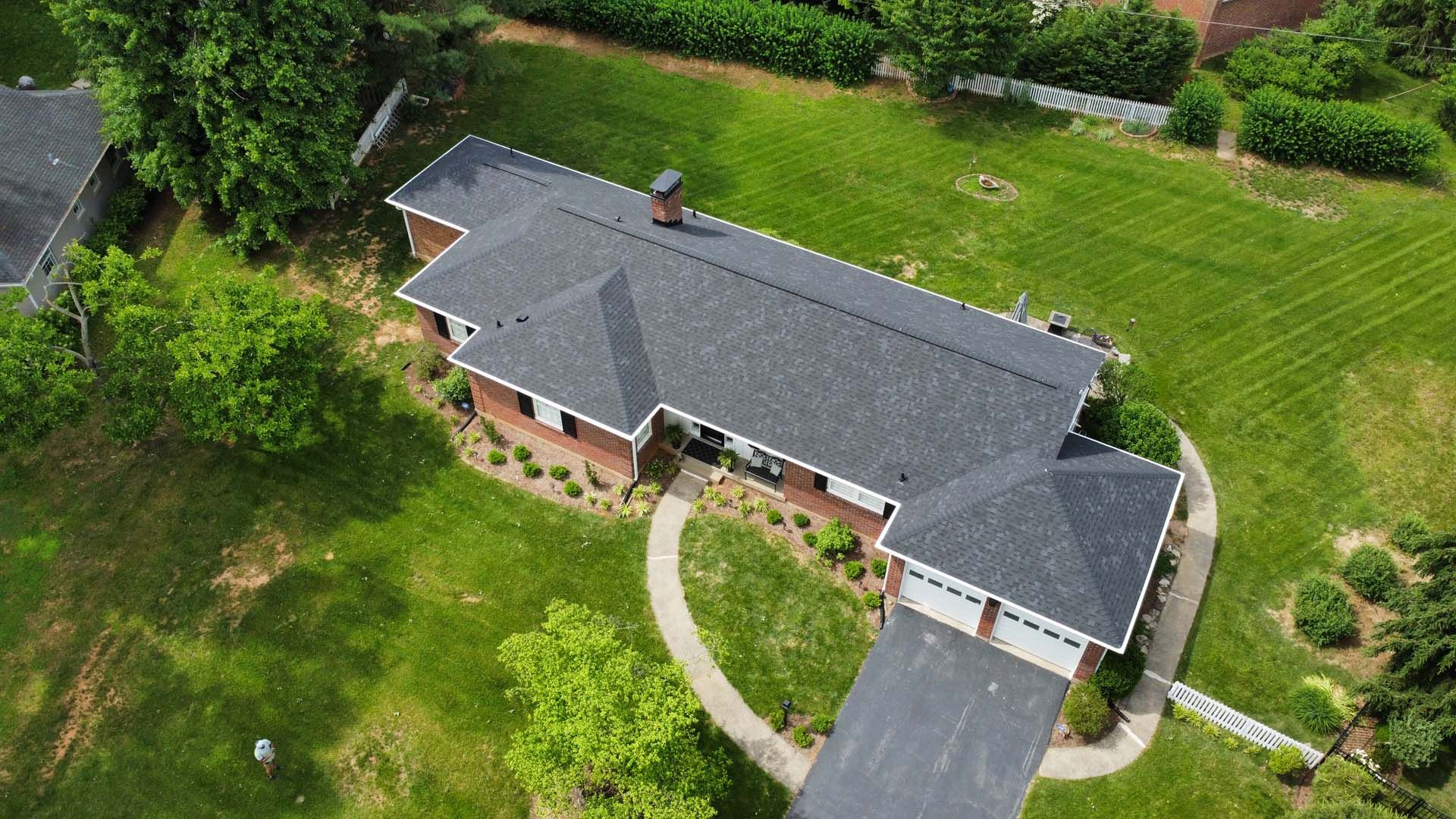 An aerial view of a single-story brick house with a dark shingled roof, surrounded by a green lawn and trees.