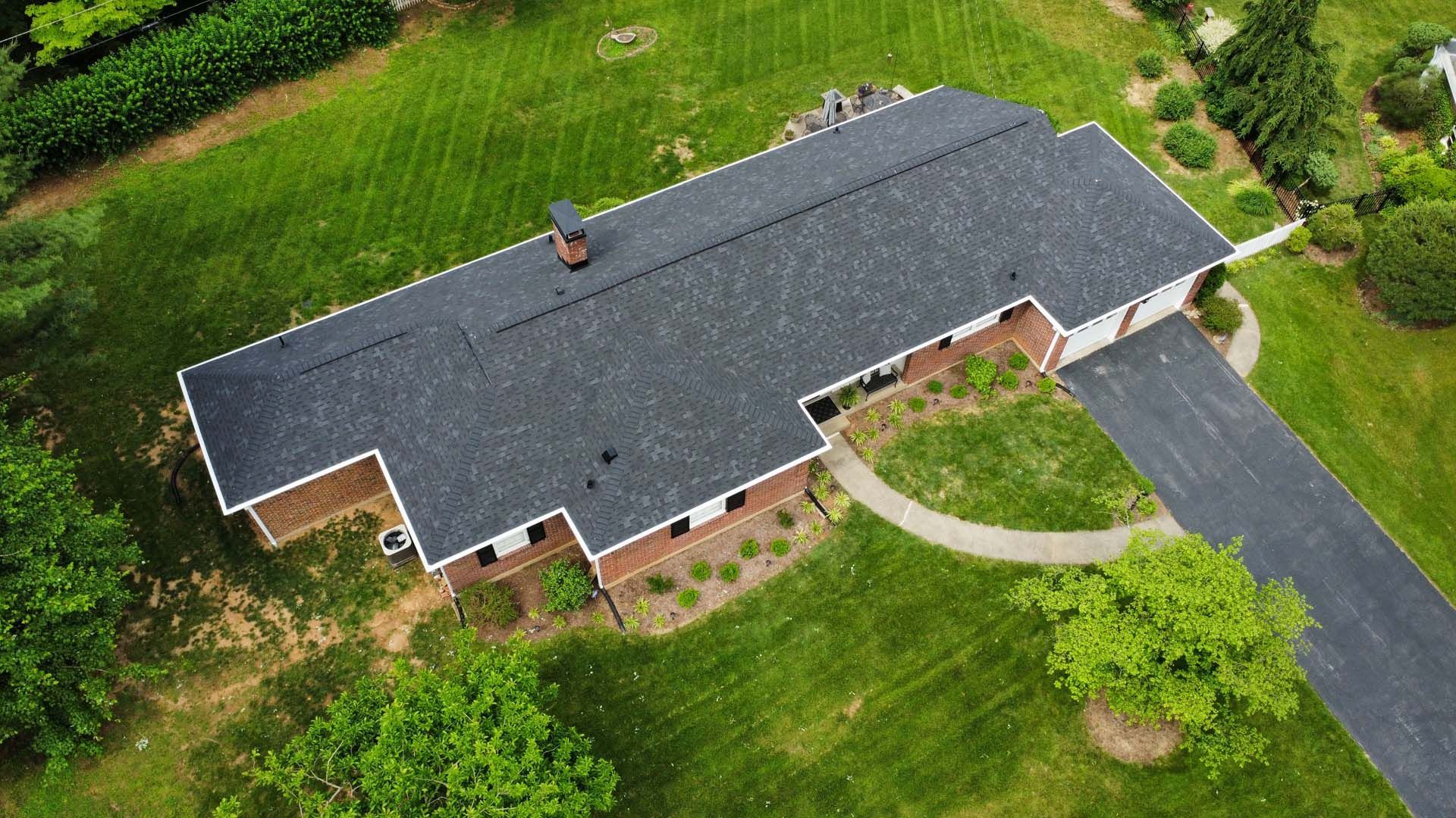 Aerial view of a single-story brick house with a dark asphalt shingle roof, surrounded by a large green lawn and driveway.