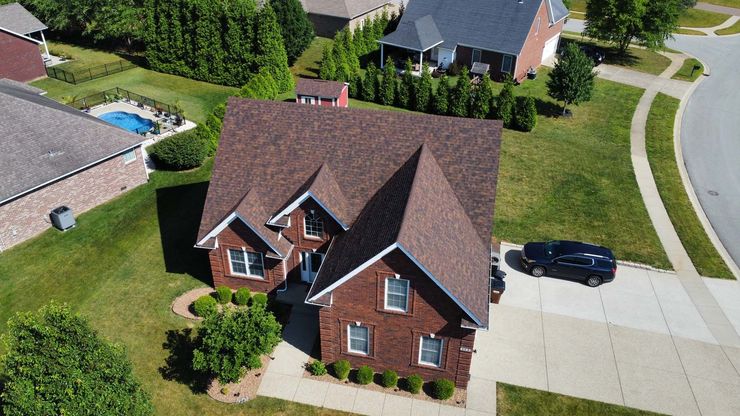 An aerial view of a multi-story, red brick house with a dark shingled roof, a driveway, and landscaped front yard.