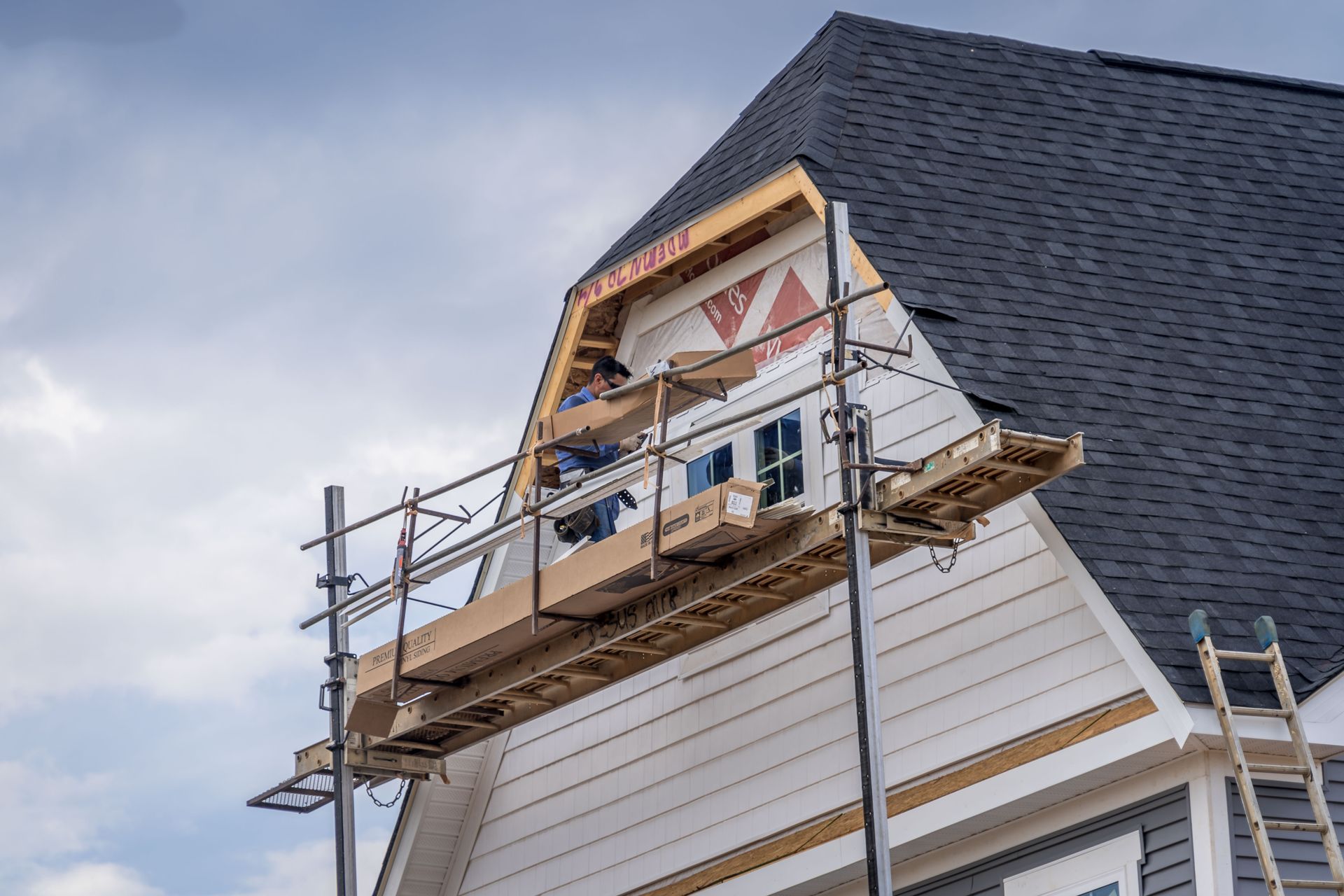 A worker on scaffolding installs siding on the upper gable of a house with a dark shingled roof under a cloudy sky.