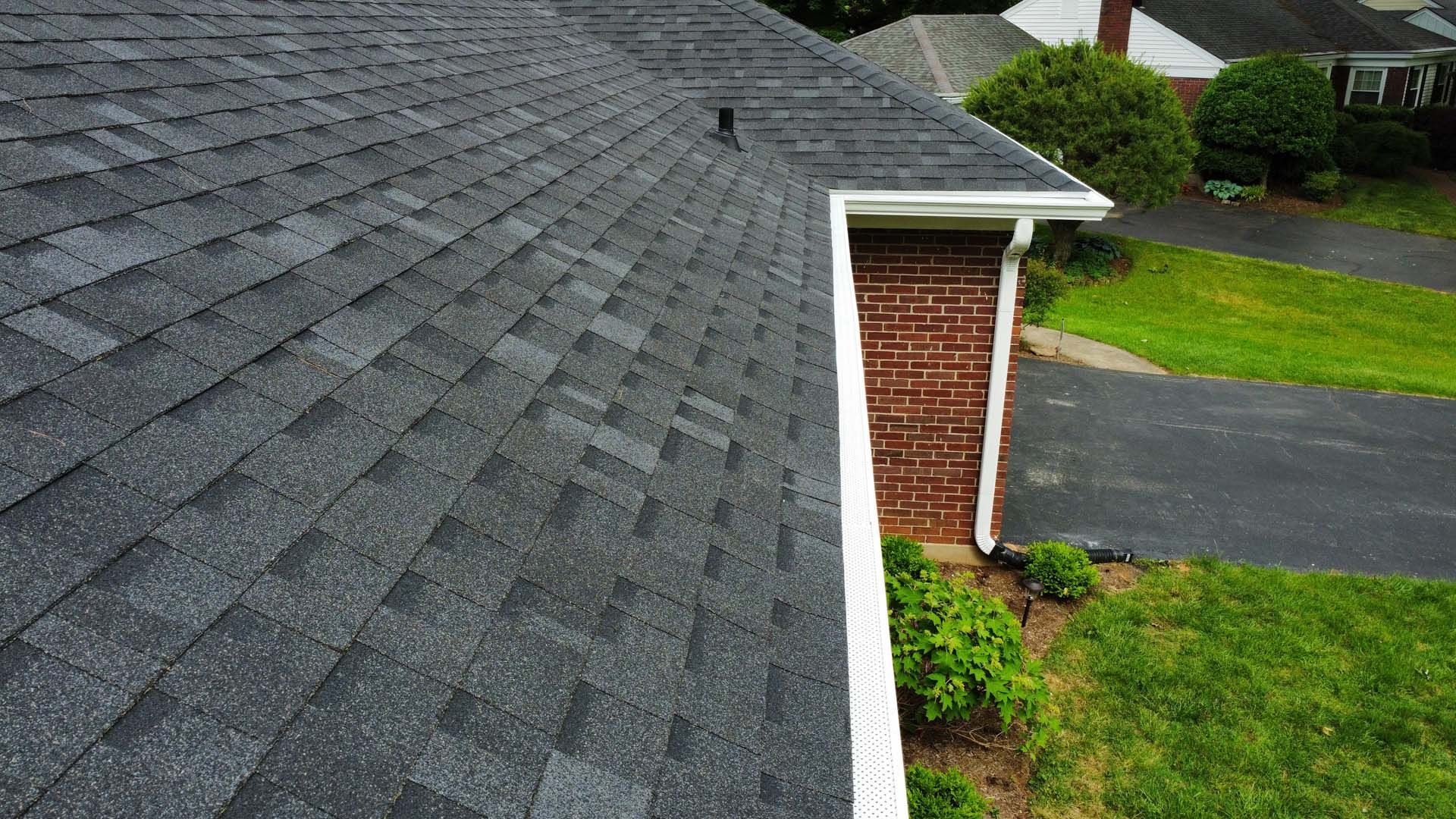 A high-angle view of a gray asphalt shingle roof, showing a corner section, white gutters, and a brick wall below.