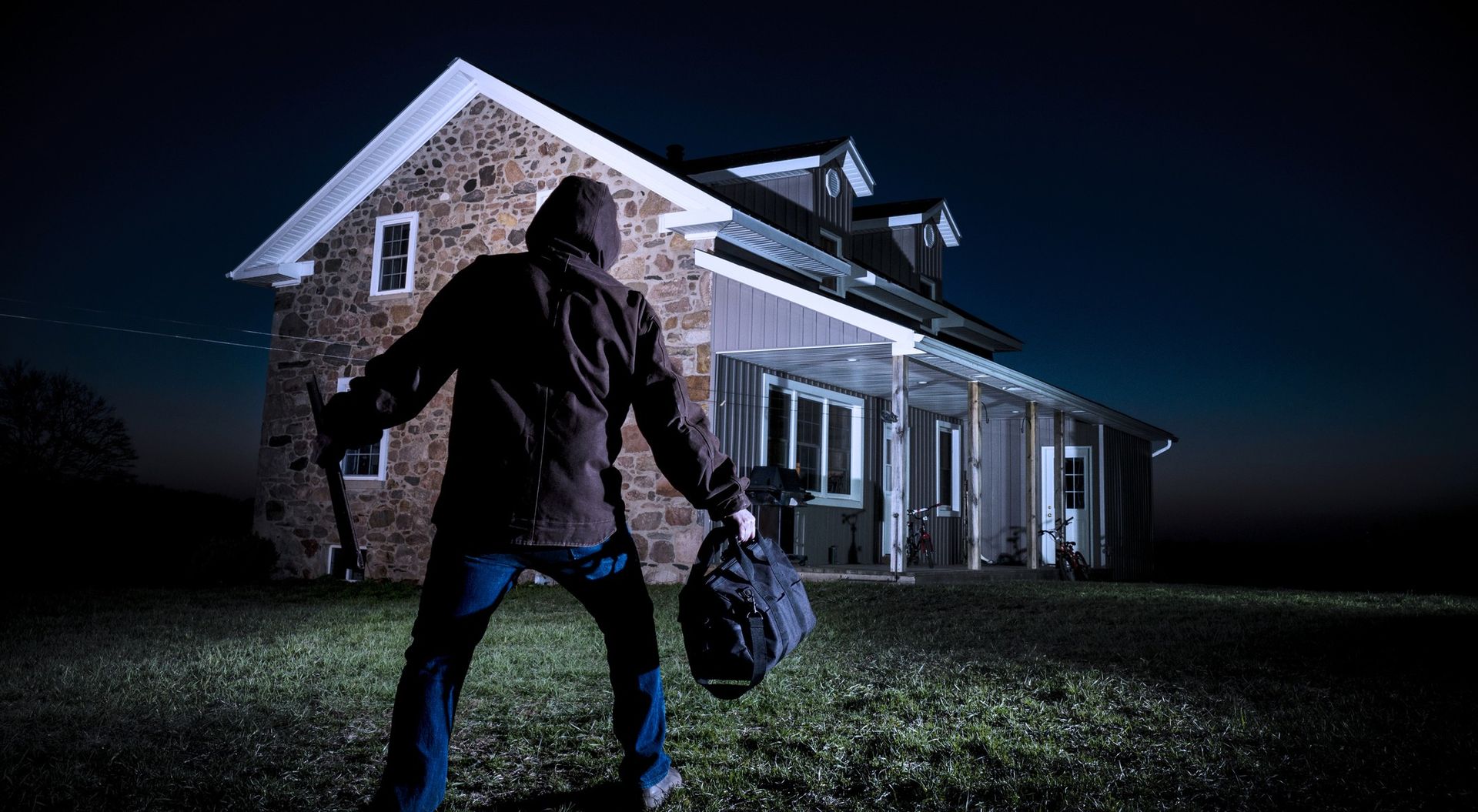 A burglar in a hooded jacket and holding a bag and crowbar stands in front of a house at night.