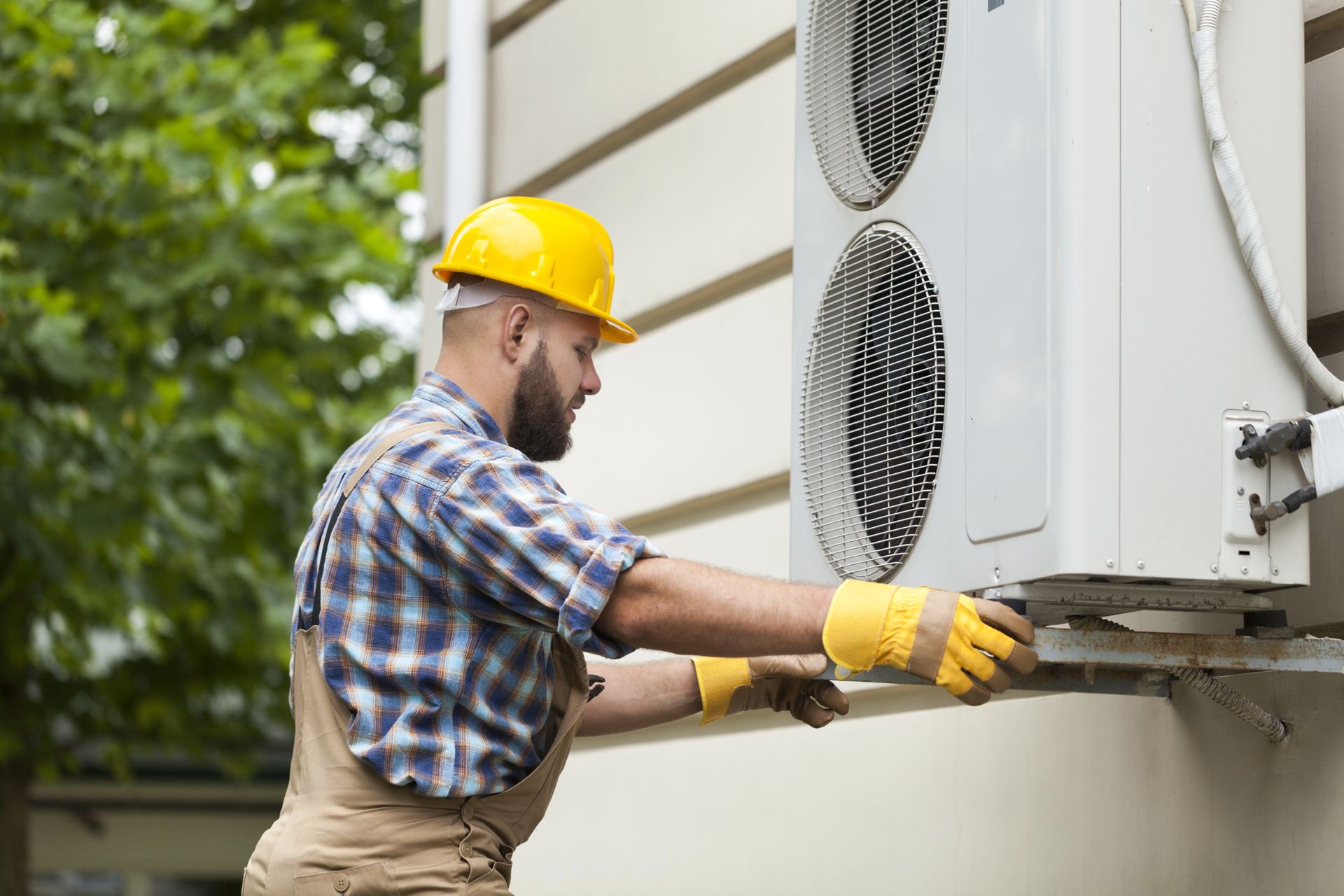 Technician in yellow hard hat and gloves repairing an outdoor air conditioning unit.