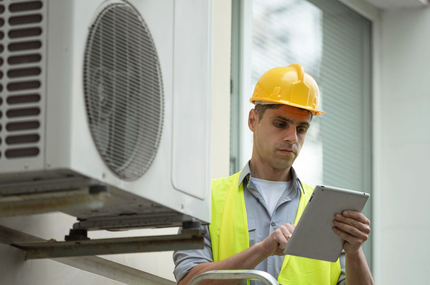 Technician in hard hat and safety vest inspecting an outdoor air conditioning unit with a tablet