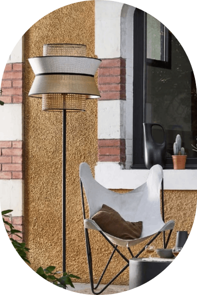 Terrashoekje met een hoge staande lamp, een witte loungestoel en een potplant bij een raam en een bakstenen muur.