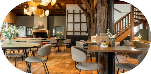 Gezellig, rustiek café-interieur met houten balken, ronde tafels, grijze stoelen en warme verlichting.