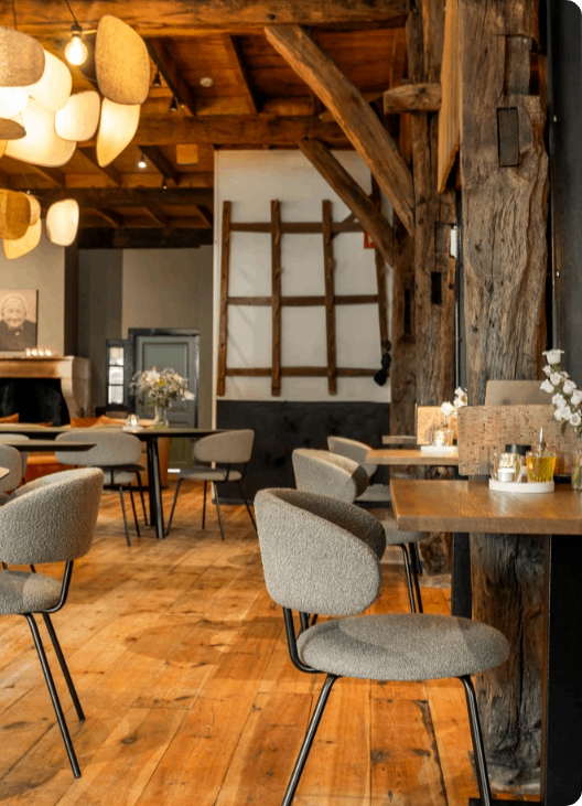Gezellig, rustiek restaurantinterieur met houten balken, ronde tafels en gestoffeerde stoelen.