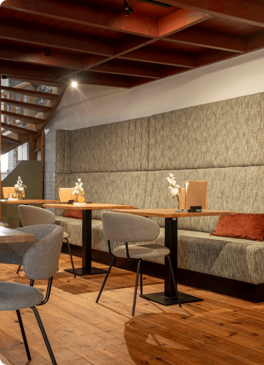 Modern café-interieur met houten tafels, gestoffeerde zitbanken en warme houten accenten aan het plafond.