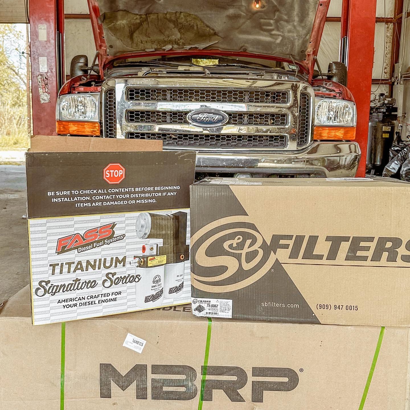 Red truck in a shop behind stacked MBRP, Synergy, and Titanium Series exhaust boxes | Proving Ground Automotive