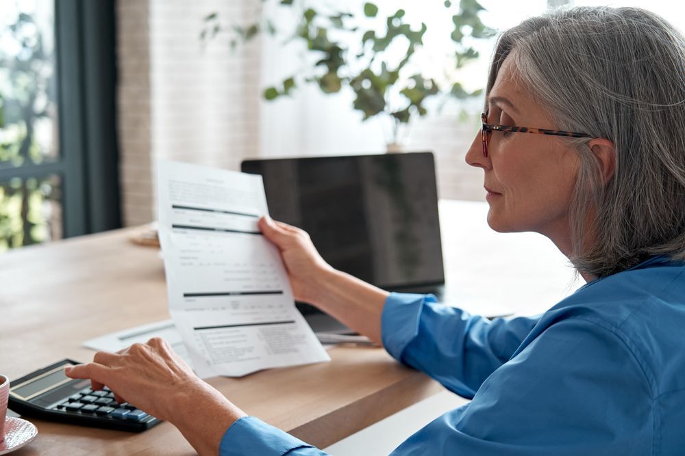 A person in a blue shirt uses a calculator while reviewing a document at a desk with a laptop.