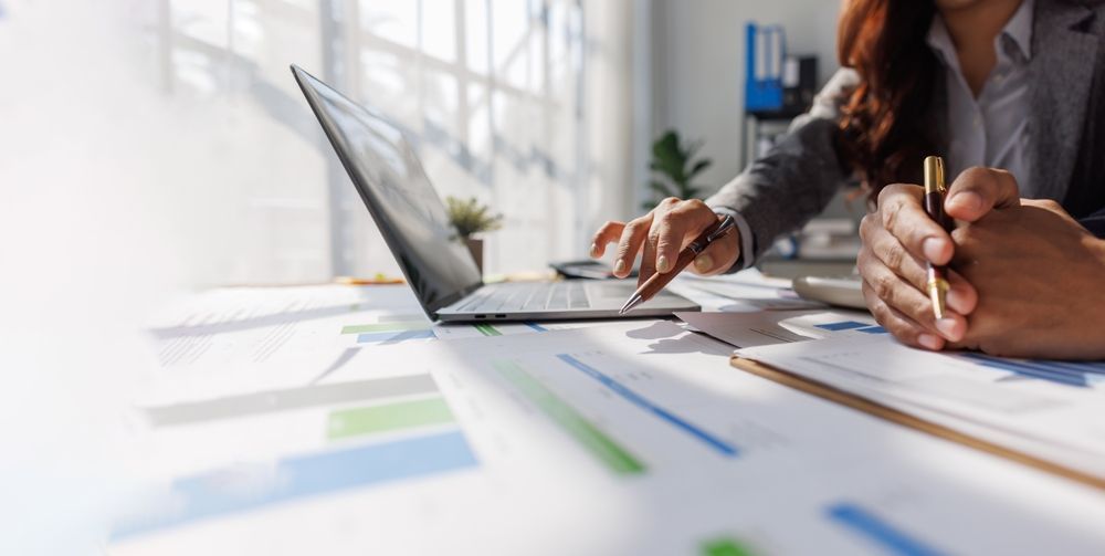 Hands and pens on a desk with financial charts and a laptop in a bright office.