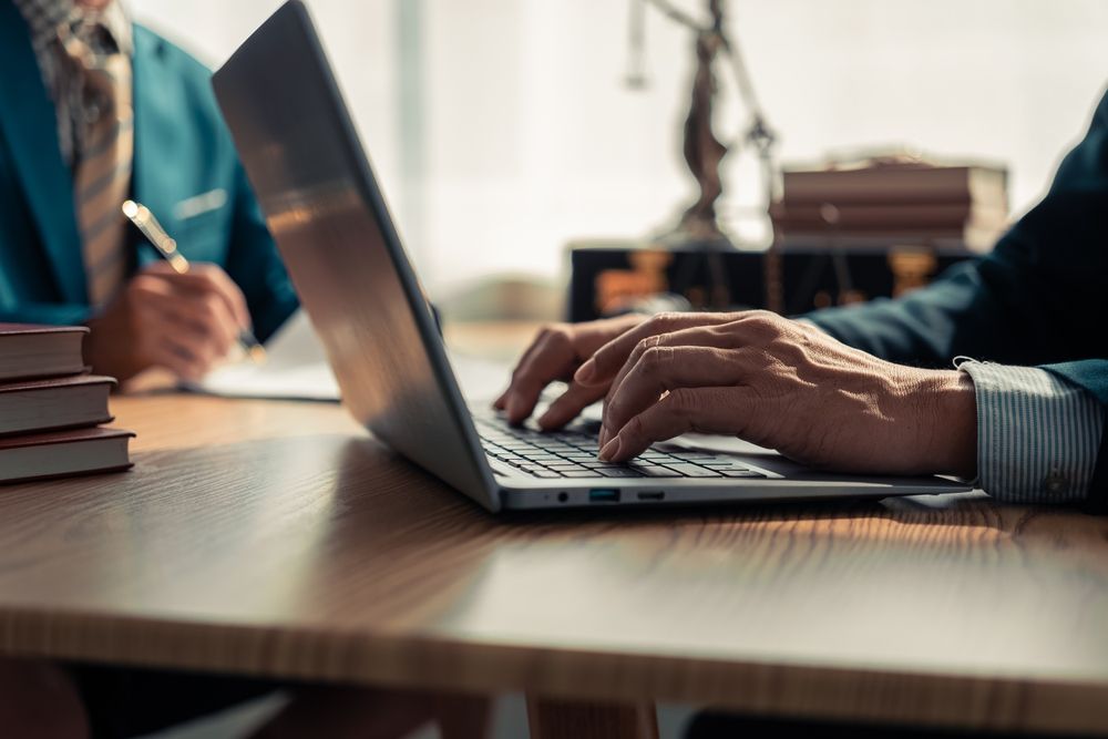 Two people in business attire work at a wooden desk with a laptop and a stack of books in an office setting.