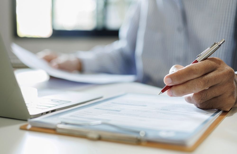 A person sitting at a desk with a laptop, using a red pen to fill out a document on a clipboard.