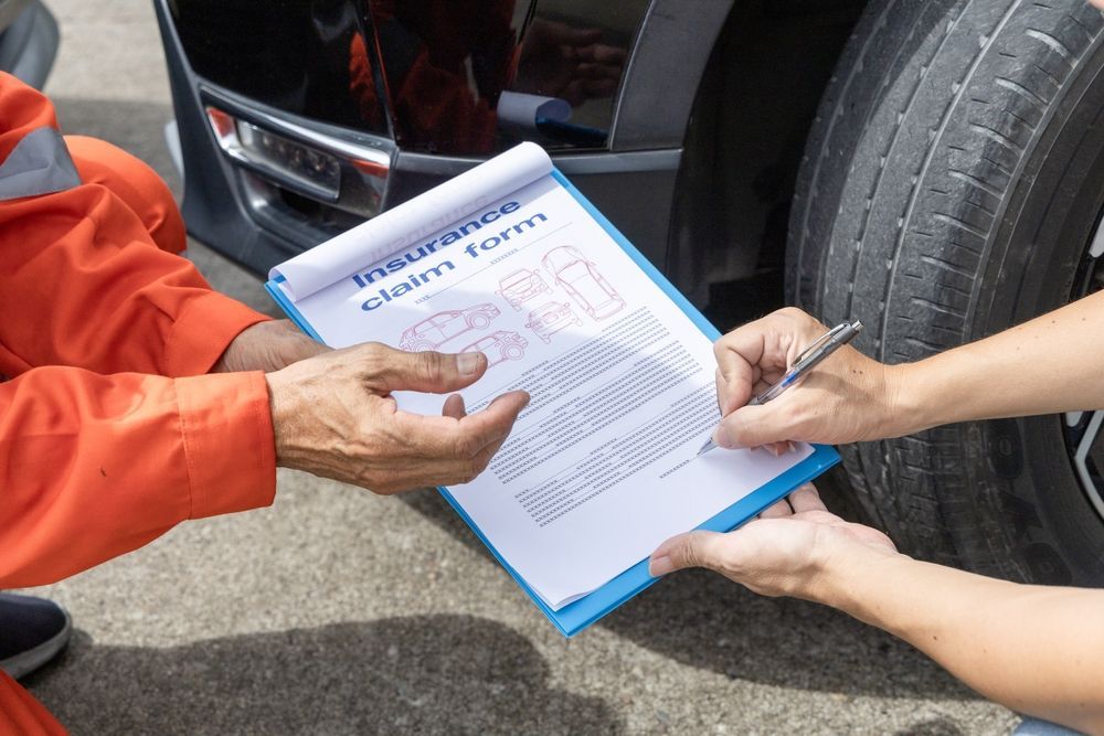 An insurance claim form is being filled out near a vehicle, with one person holding the clipboard and another writing.