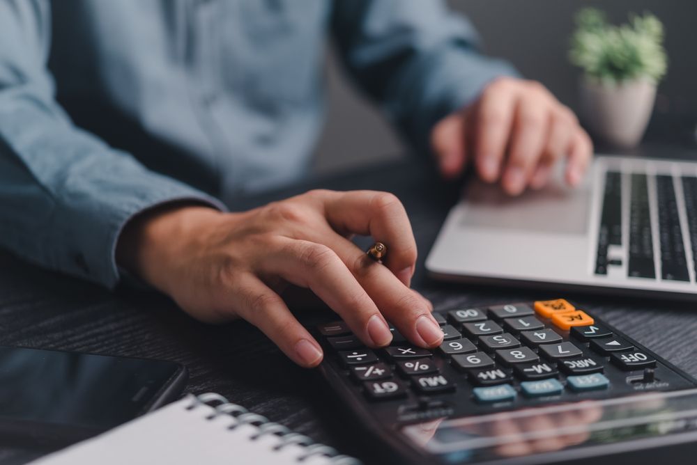 A person in a blue shirt uses a calculator on a desk next to a laptop and notebook.
