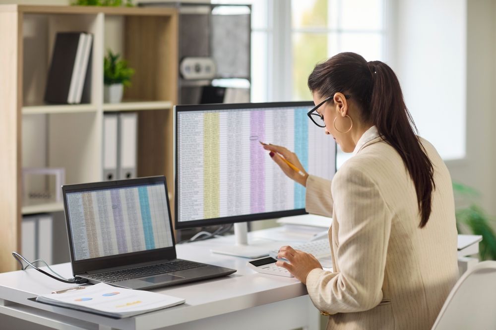 A professional sitting at a desk, pointing at a computer monitor displaying a spreadsheet while using a calculator.