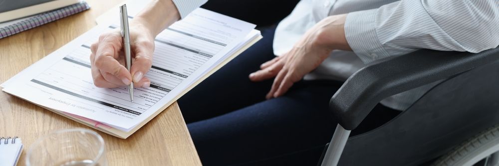 A person in a wheelchair writing on a document at a wooden desk.