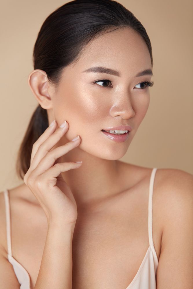 Woman with a light complexion, touching her cheek, against a light beige background.