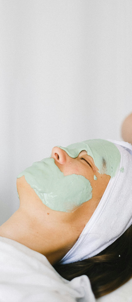 Person with a green facial mask, resting on a white towel, in a spa setting.