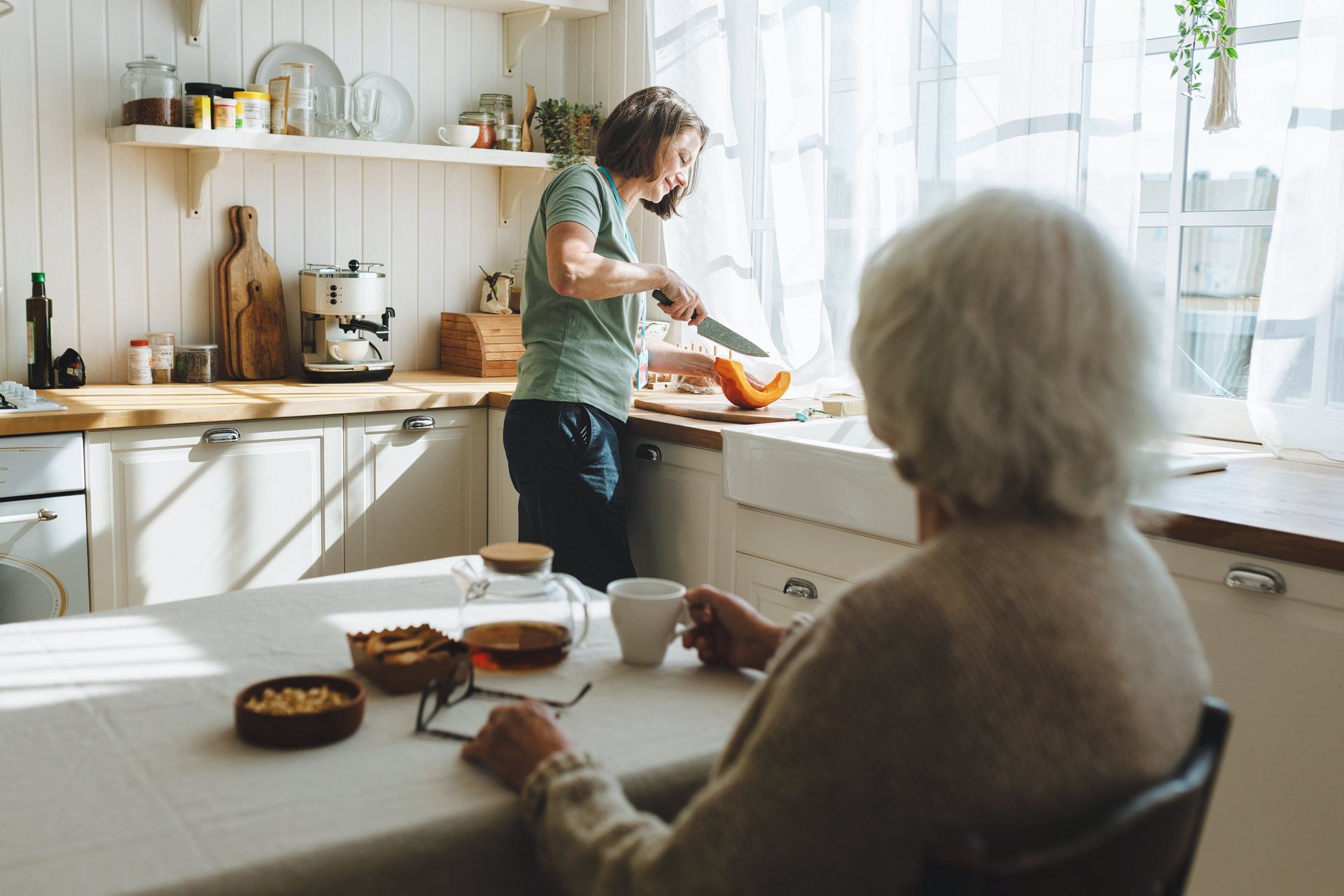 One person prepares food at a kitchen counter while another person sits at a table in the foreground.