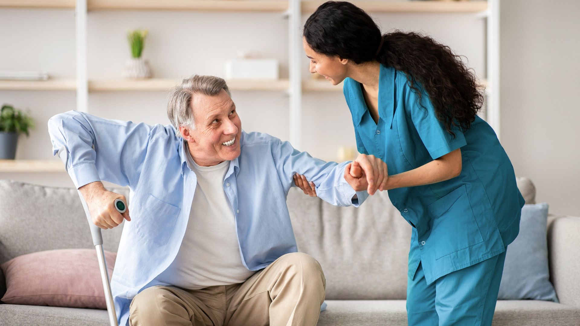 A healthcare provider in blue scrubs assists a smiling person with a cane as they stand up from a couch in a living room.