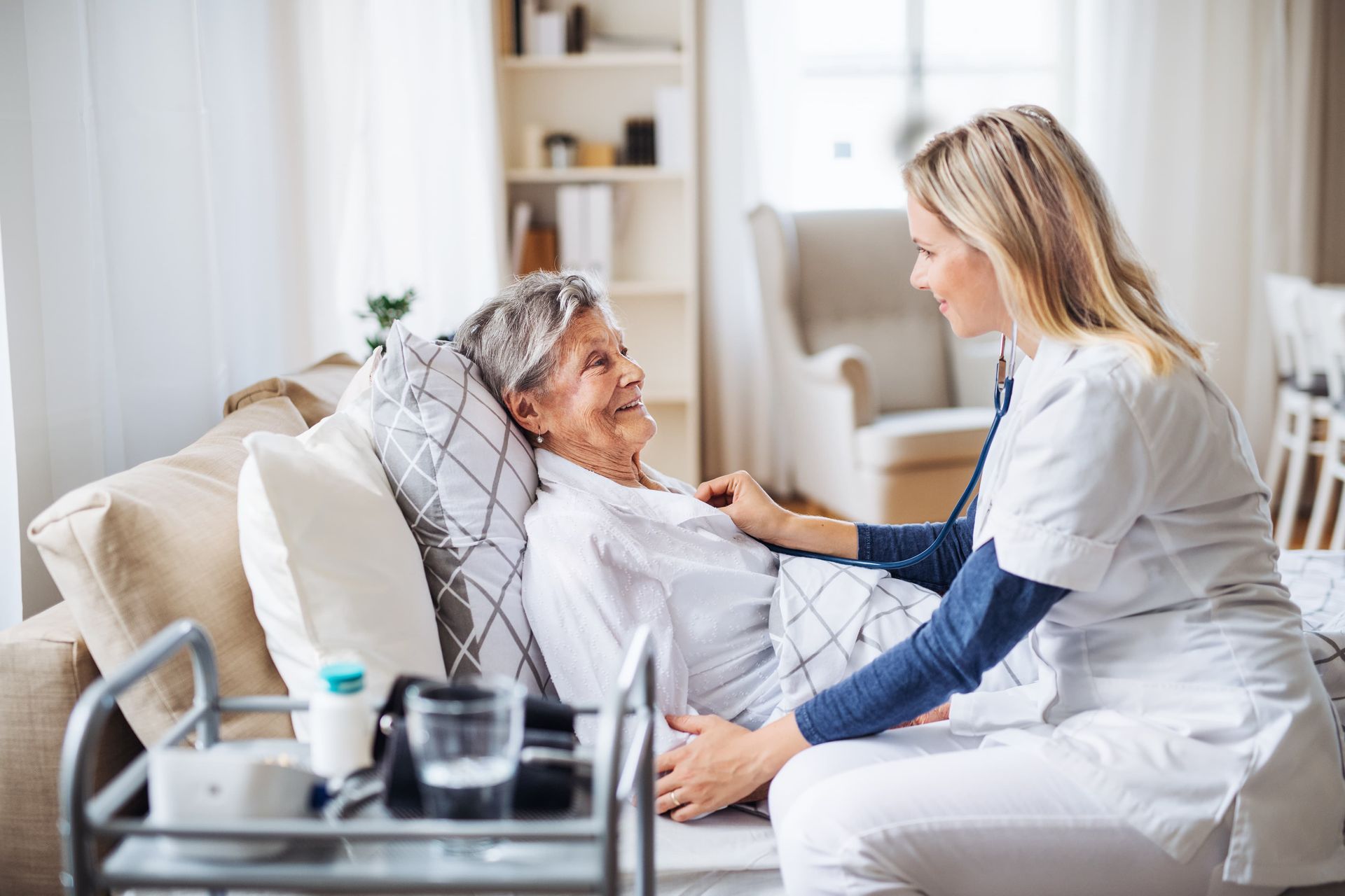 A home healthcare professional uses a stethoscope to examine an elderly patient resting on a couch.