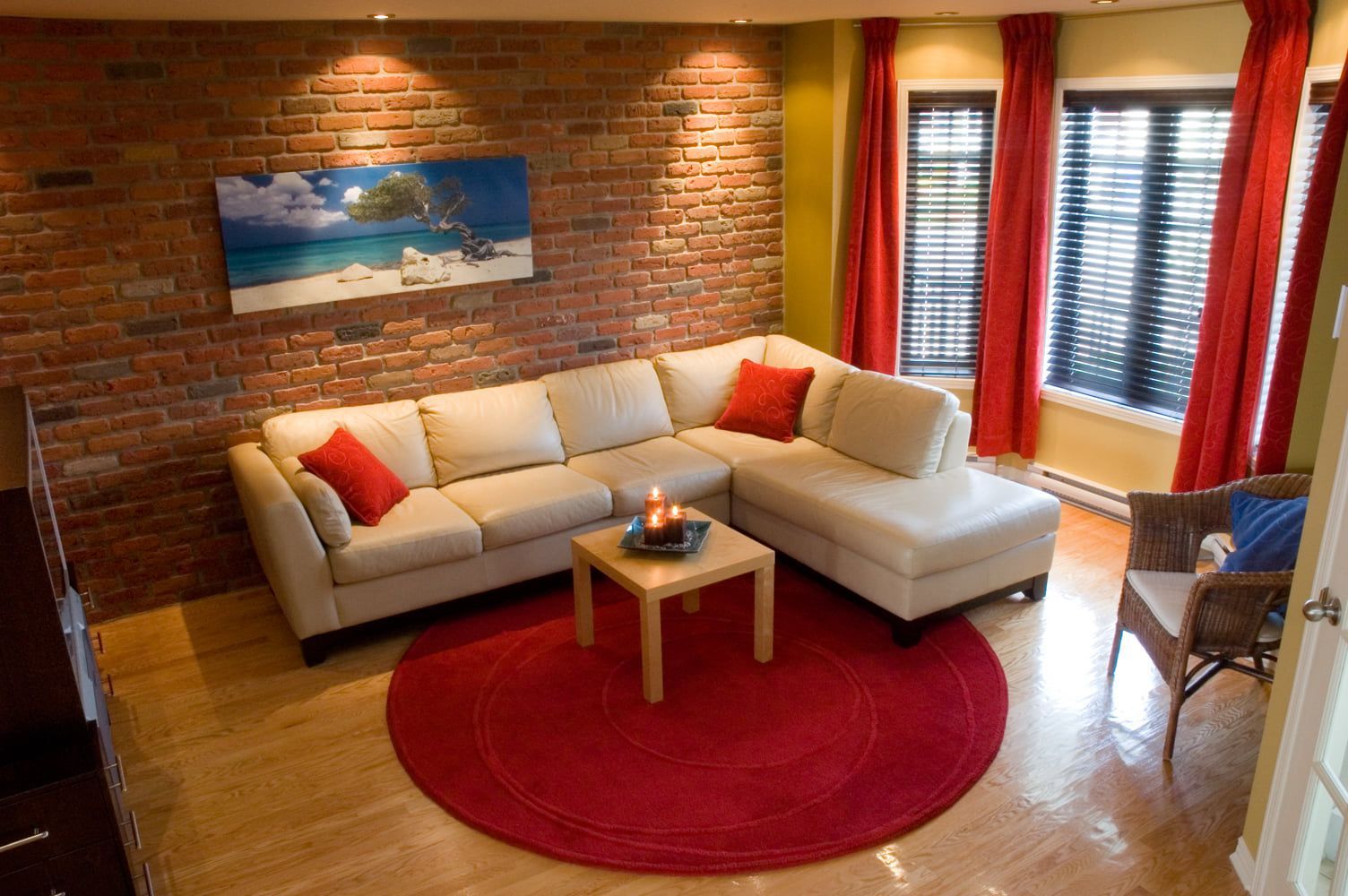 Salon avec canapé d'angle beige, tapis rouge, mur de briques, œuvres d'art et rideaux rouges.
