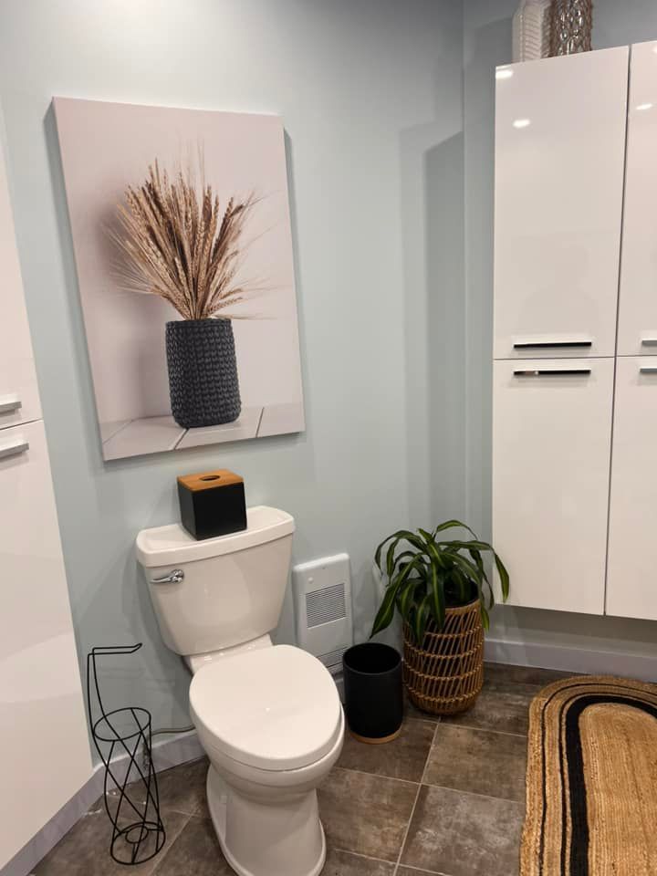 Salle de bains avec toilettes, tableau représentant un vase rempli de blé et armoires de rangement blanches.