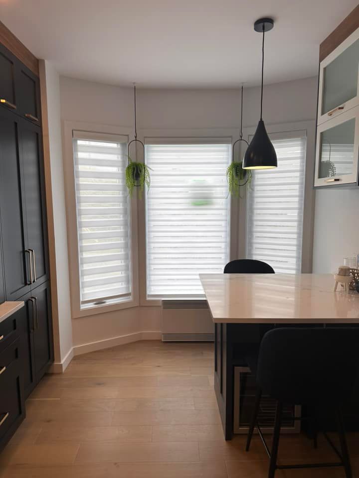 Cuisine avec parquet clair, stores blancs et touches de noir. Des plantes suspendues ornent le devant de la fenêtre.