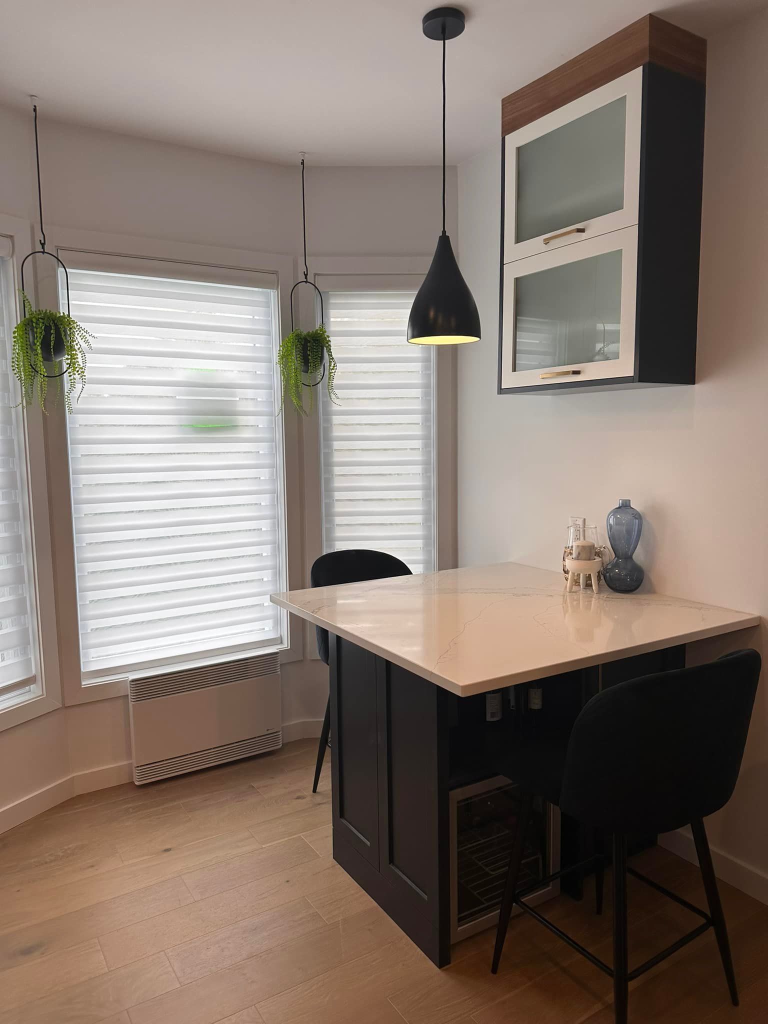 Cuisine avec un îlot central foncé et un plan de travail blanc, deux chaises noires, une suspension lumineuse et des plantes.