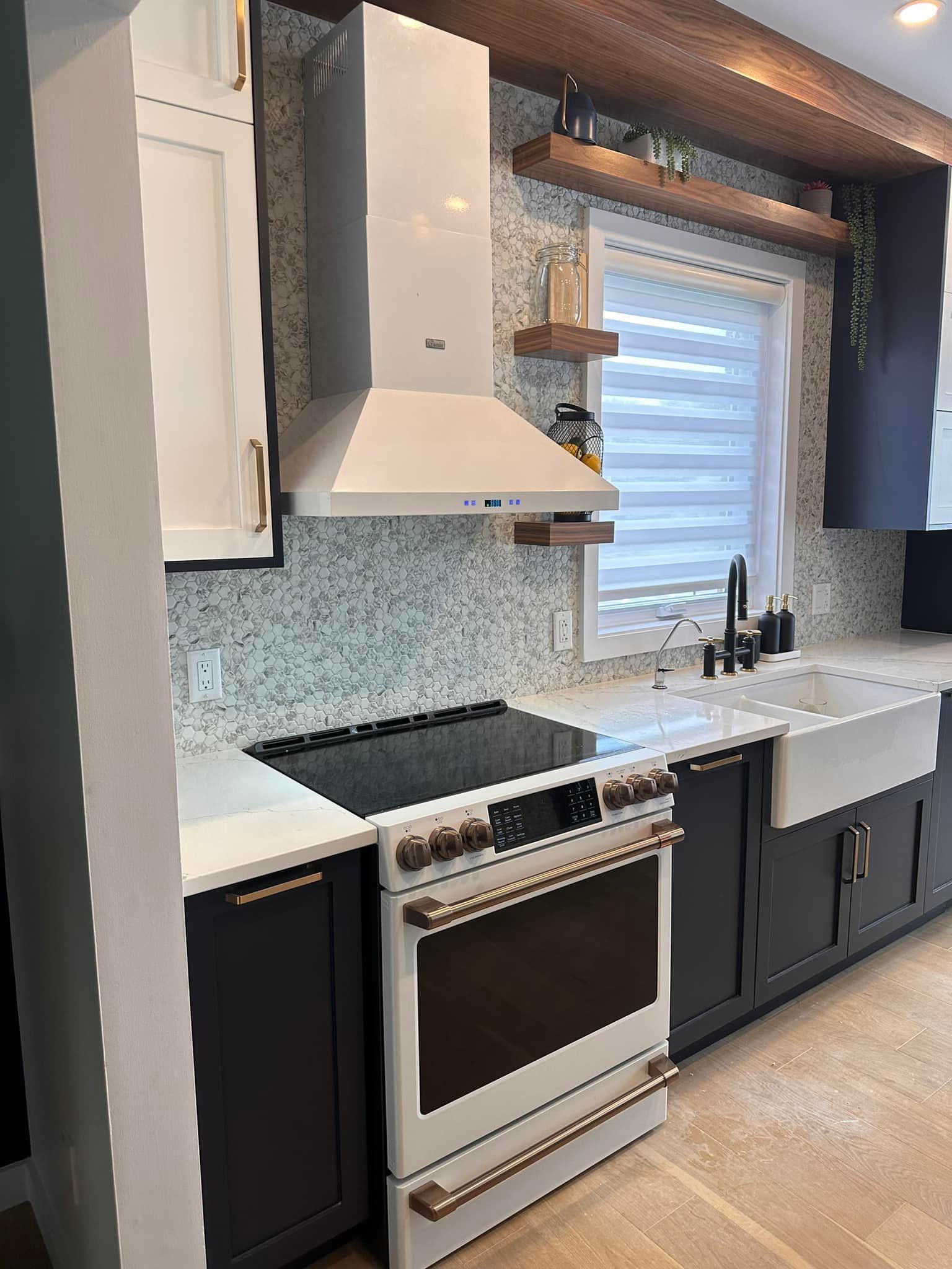 Cuisine moderne avec armoires blanches et foncées, crédence claire, cuisinière et hotte aspirante, évier de style campagnard et étagères en bois.