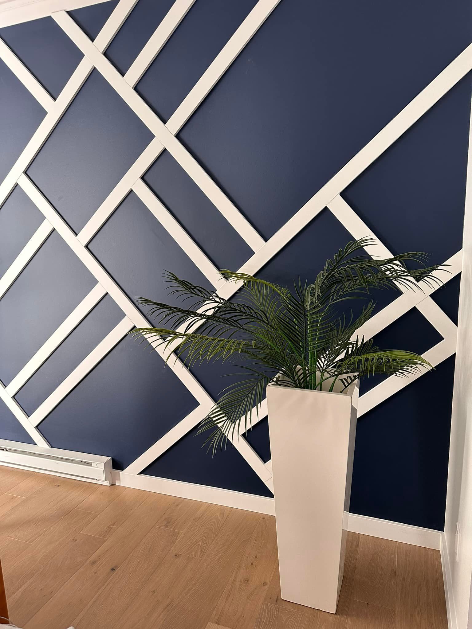 Mur bleu orné de moulures géométriques blanches, parquet et grand pot de fleurs blanc rempli de verdure.