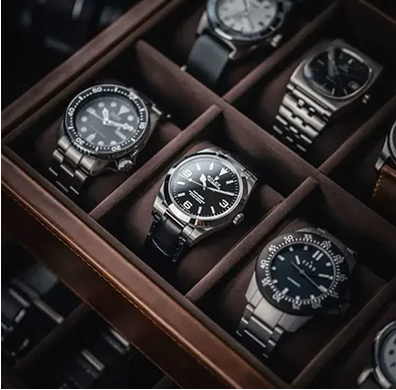 A high-angle close-up of a brown wooden watch box displaying a collection of various stainless steel wristwatches.