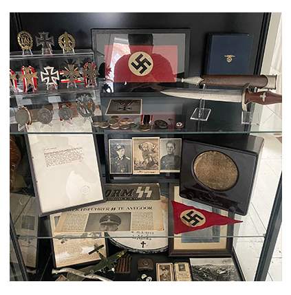 A glass display cabinet filled with historical Nazi-era militaria, medals, daggers, photographs, and printed materials.