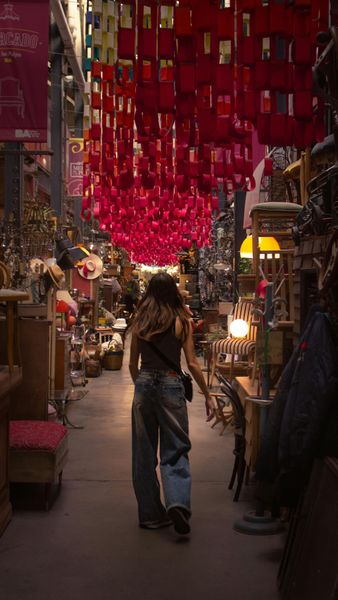 A person walks down an aisle in an antique shop filled with vintage items, under a ceiling covered in hanging red fabric.