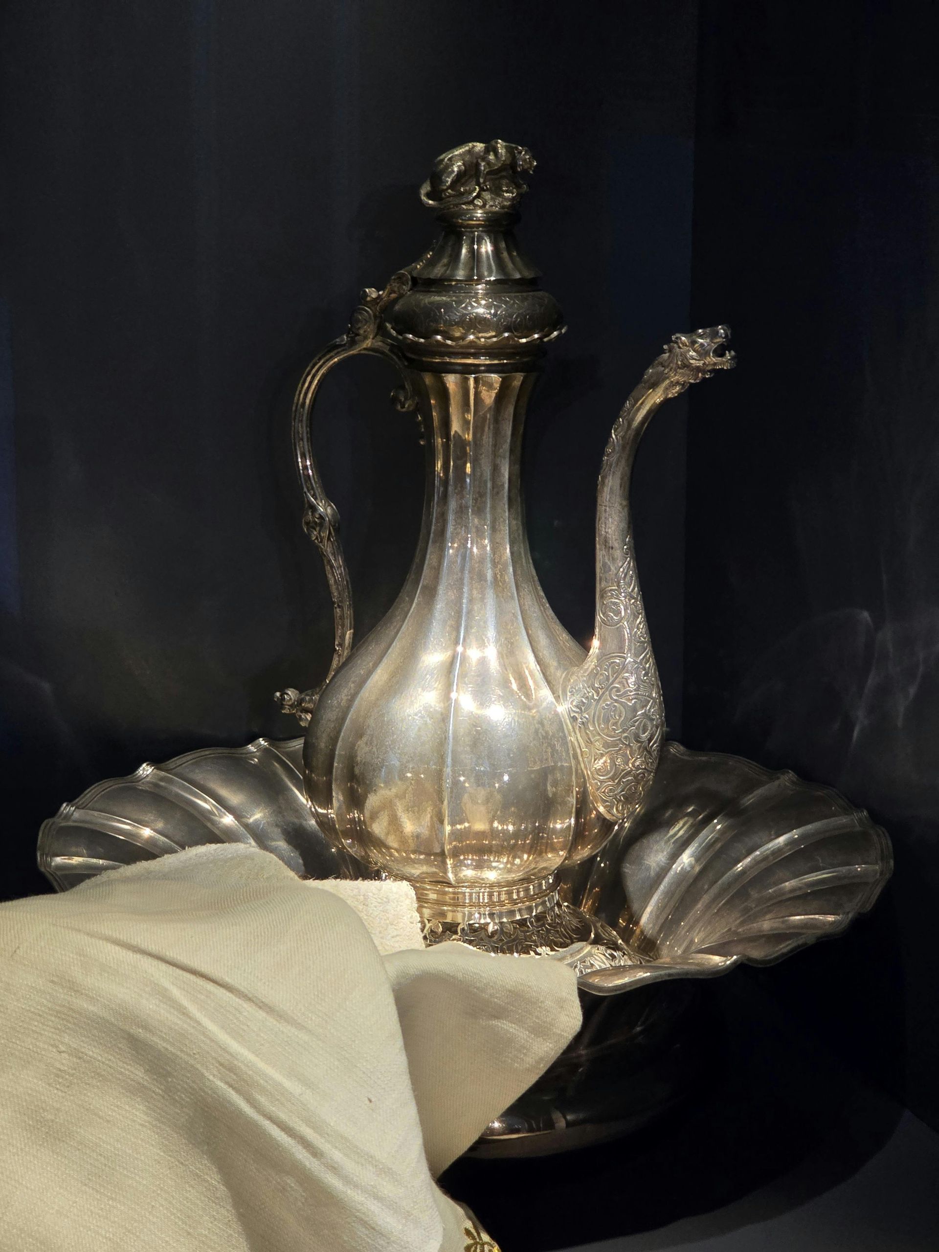 An ornate silver ewer and basin set with a patterned cloth draped in the foreground against a dark background.