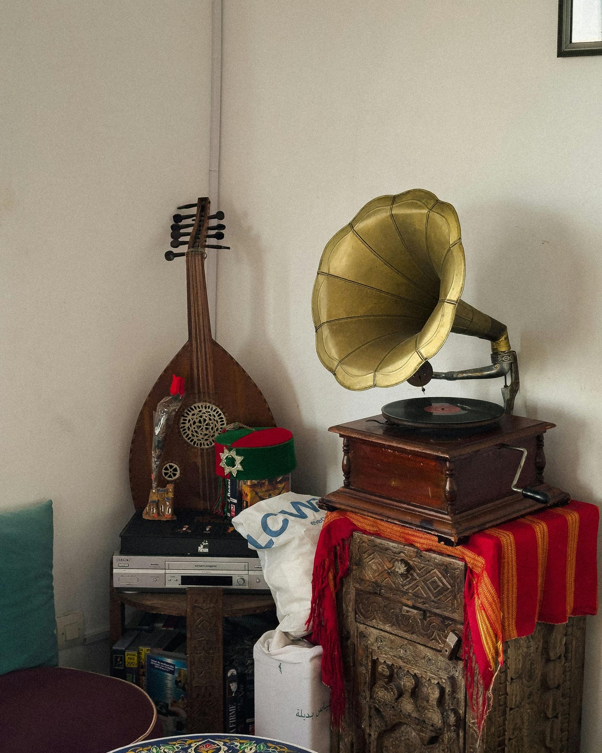 A vintage gramophone sits on a carved wooden cabinet beside an oud instrument in the corner of a room.