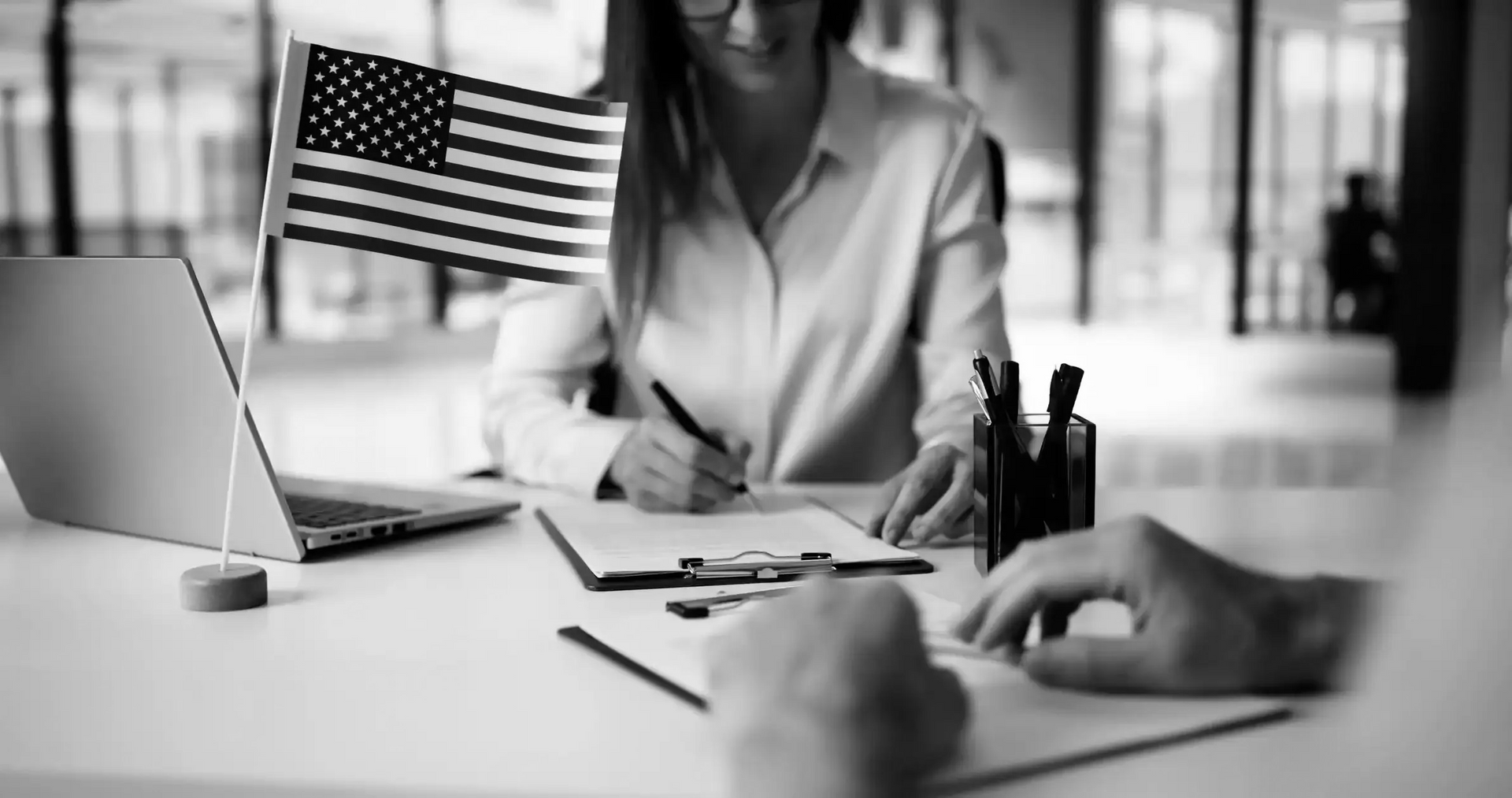 Woman signing documents at a desk, US flag, laptop, hands, and pens.