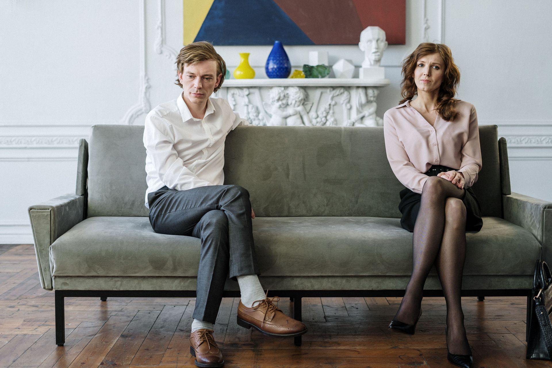 Two people seated on a gray sofa in a bright living room, with colorful vases on the shelf behind them.