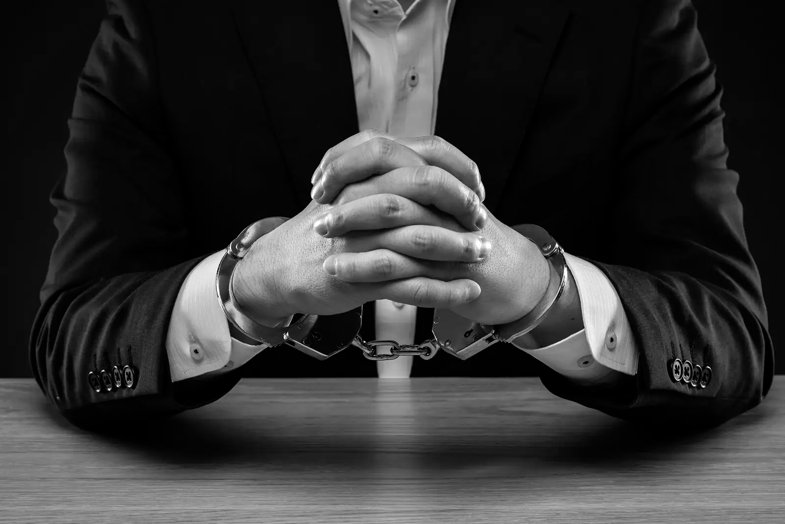 Man in suit, hands clasped, wearing handcuffs at a table.