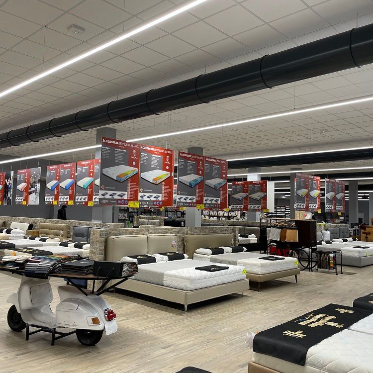 Interieur van een matrassenwinkel met veel bedden in de vitrine, plafondverlichting, metalen geperforeere luchttoevoerkanalen en reclameborden.